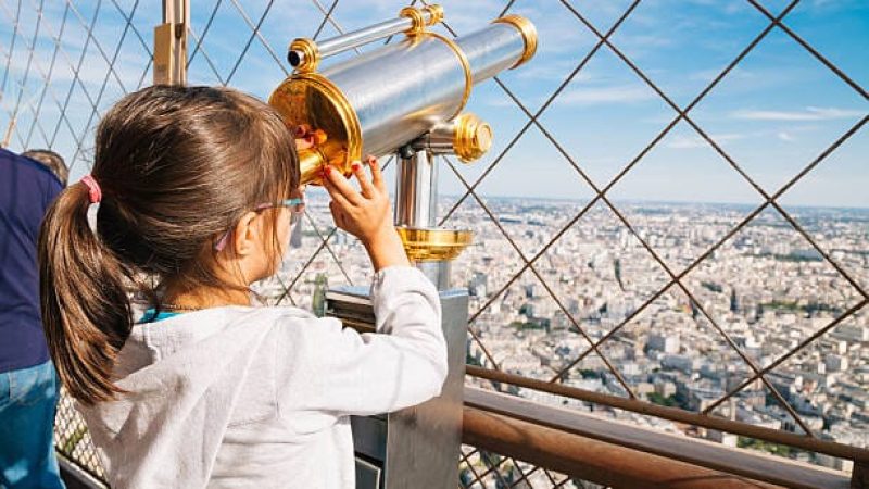 Little girl looking the cityscape of Paris from the top floor of the Eiffel Tower using the telescope